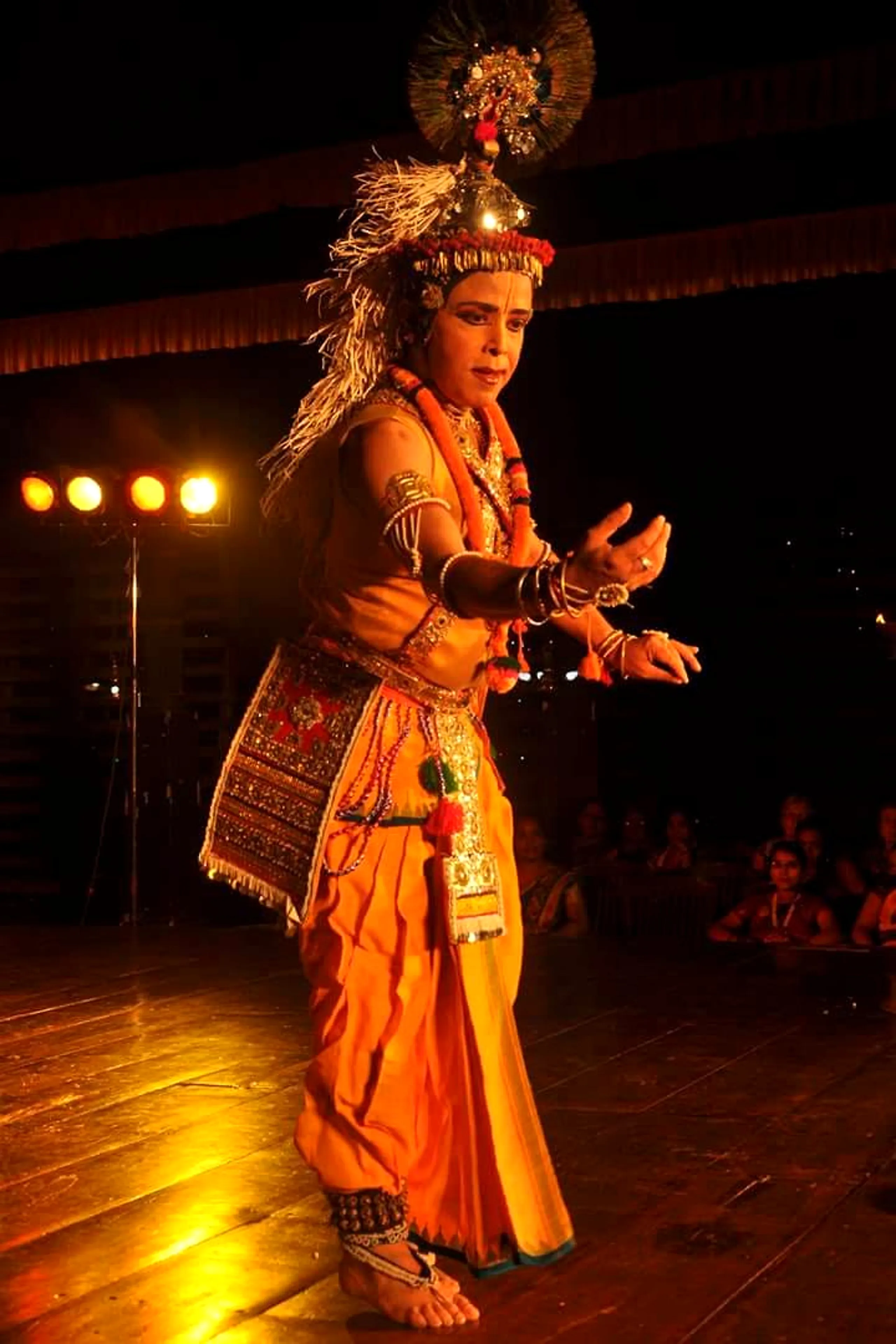 Sanjib in saffron costume and ornate headdress, spotlit mid-gesture on a darkened stage